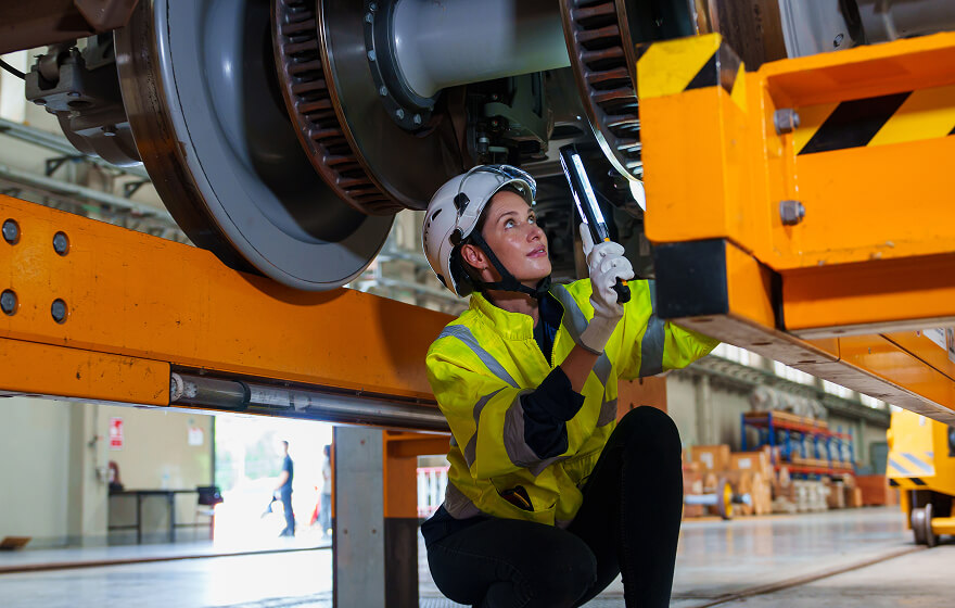 A woman wearing safety gear crouching under a railway to work on repair.